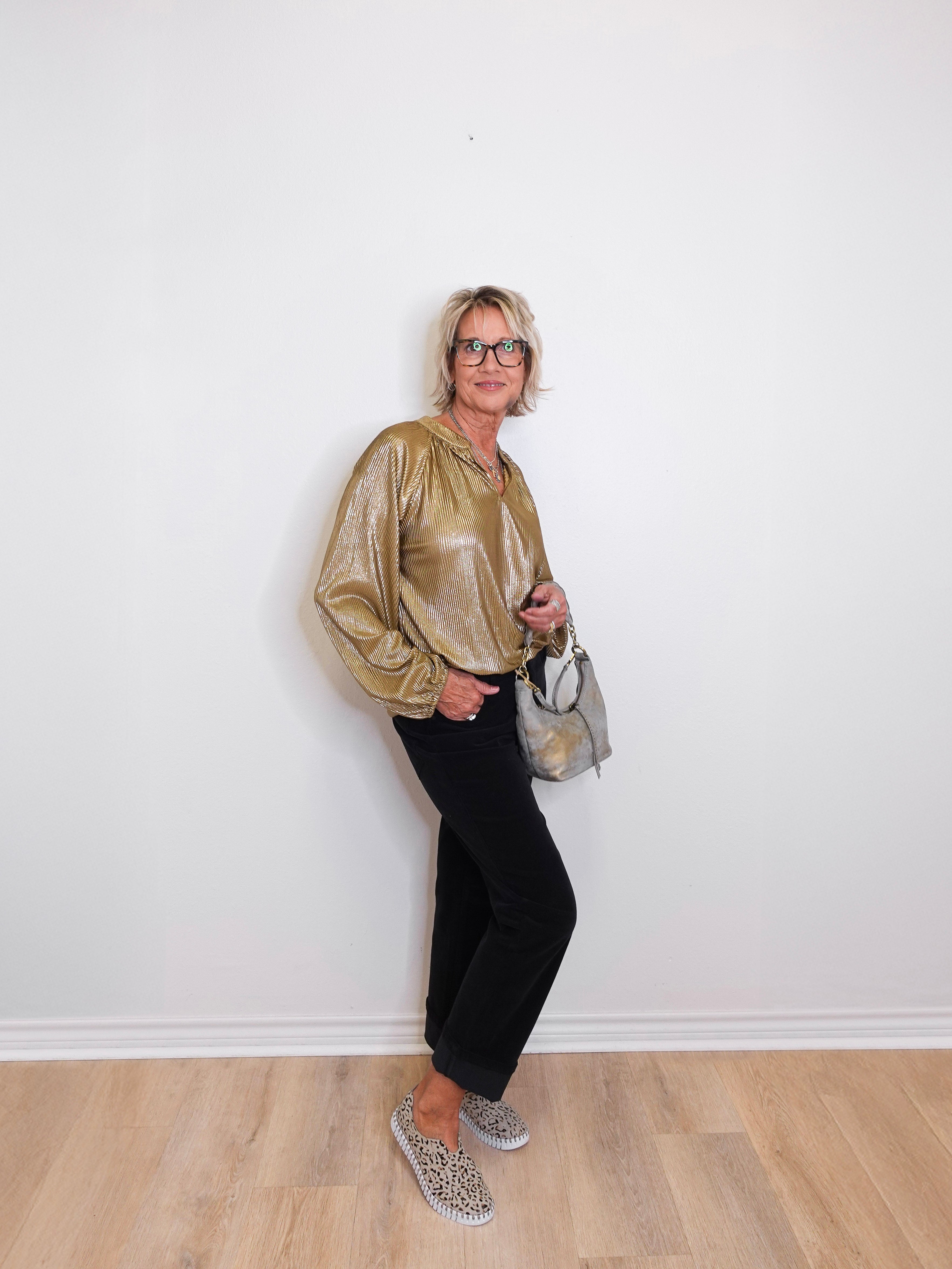 Woman wearing a gold blouse, black pants, and silver shoes against a white wall.