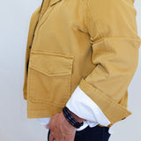 A person wearing a mustard yellow utility jacket over a white collared shirt and dark jeans, shown from the chest down against a plain light wall.