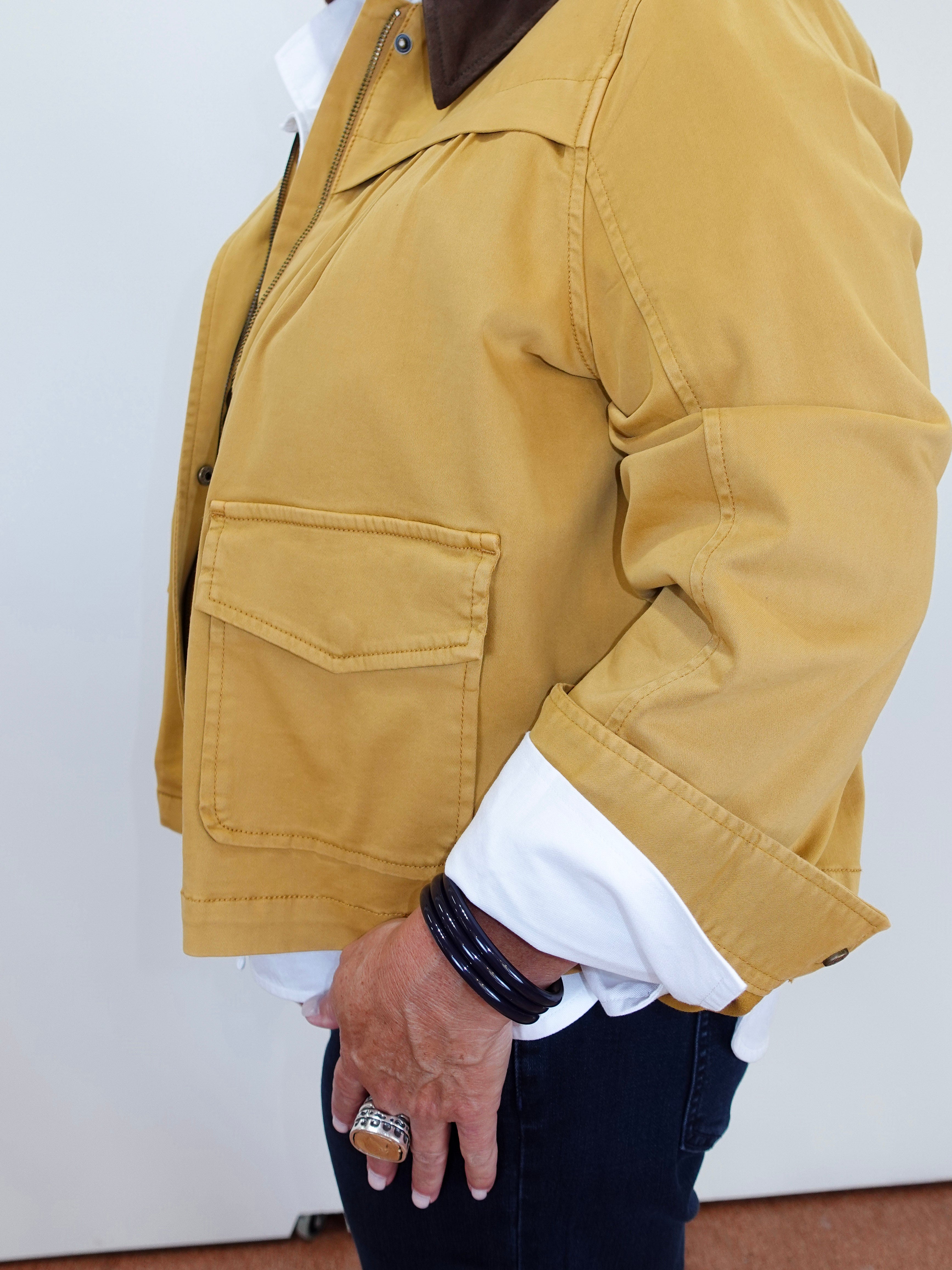 A person wearing a mustard yellow utility jacket over a white collared shirt and dark jeans, shown from the chest down against a plain light wall.