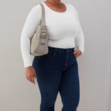 Person wearing a white long-sleeve top and blue jeans, holding a beige handbag against a plain background