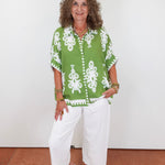 Woman wearing a green patterned shirt and white pants against a white wall.