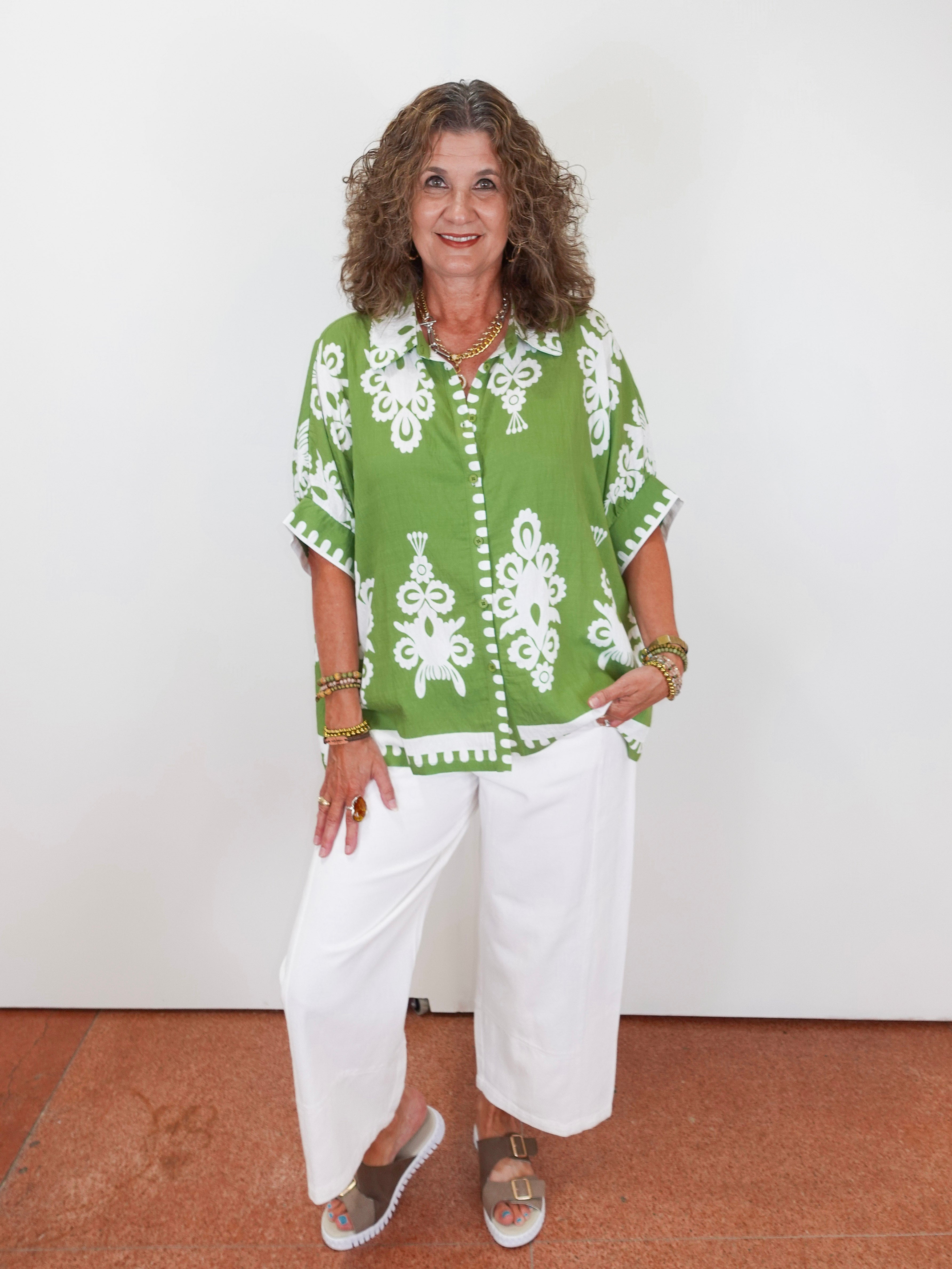 Woman wearing a green patterned shirt and white pants against a white wall.