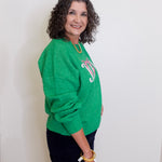 Woman wearing a green sweater with a logo, standing against a white wall.