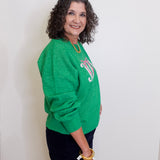 Woman wearing a green sweater with a logo, standing against a white wall.