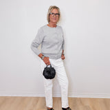 Woman wearing a gray sweater, white pants, and black shoes holding a black handbag against a white wall.