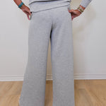 Person wearing a gray outfit with wide-leg pants against a white wall.