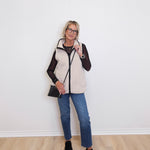 Woman wearing a white puffer vest over a black long-sleeve shirt, blue jeans, and black shoes against a white wall.