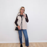 Woman wearing a white puffer vest over a black long-sleeve shirt, blue jeans, and black shoes against a white wall.