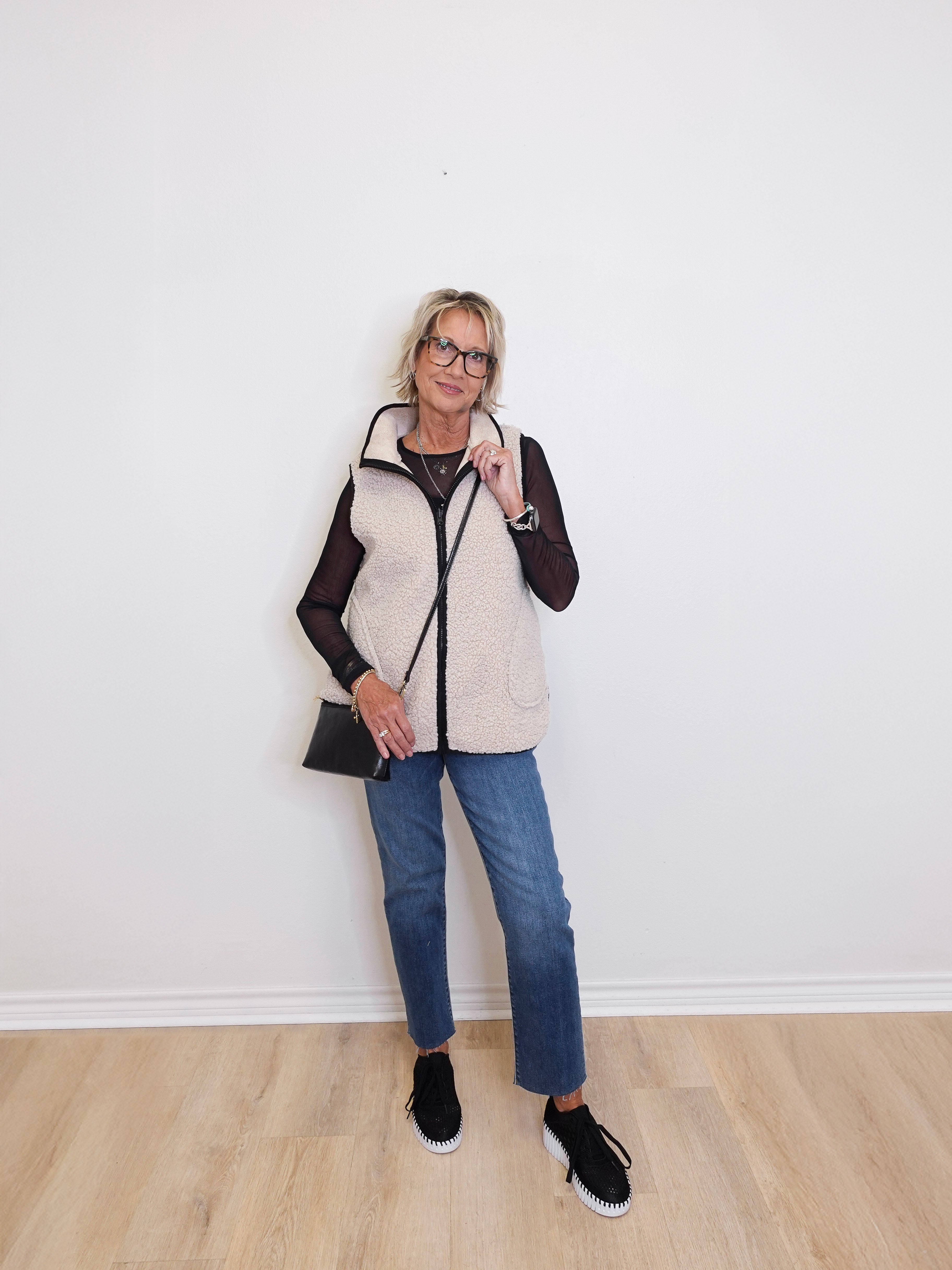 Woman wearing a white puffer vest over a black long-sleeve shirt, blue jeans, and black shoes against a white wall.