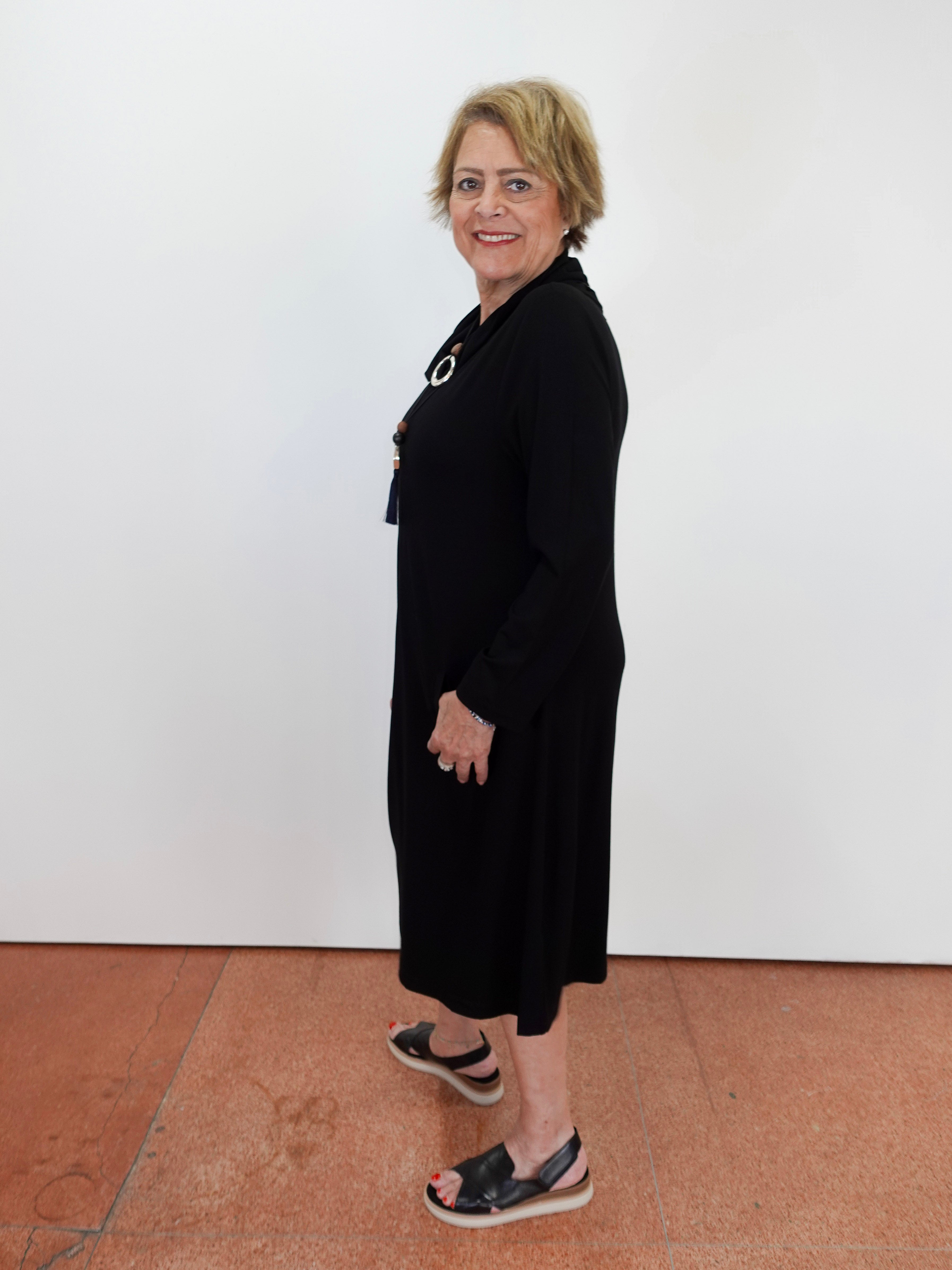 A woman with short blonde hair wearing a black long-sleeve midi dress and black slide sandals, standing against a white wall on a terracotta tiled floor.