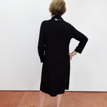A woman with short blonde hair is shown from behind wearing a black long-sleeve midi dress and black sandals, standing against a plain white wall on a terracotta-colored floor.