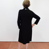 A woman with short blonde hair is shown from behind wearing a black long-sleeve midi dress and black sandals, standing against a plain white wall on a terracotta-colored floor.