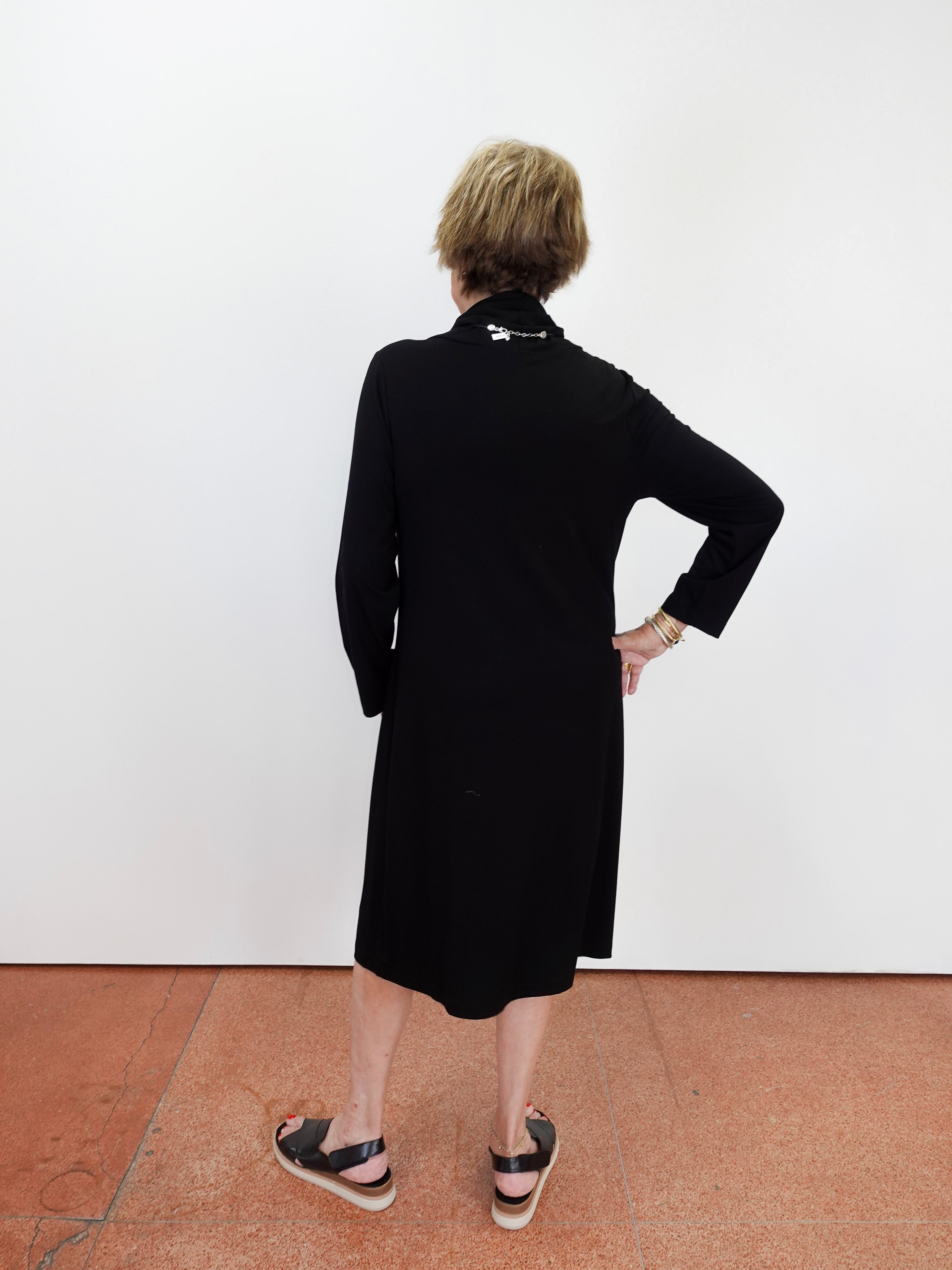 A woman with short blonde hair is shown from behind wearing a black long-sleeve midi dress and black sandals, standing against a plain white wall on a terracotta-colored floor.