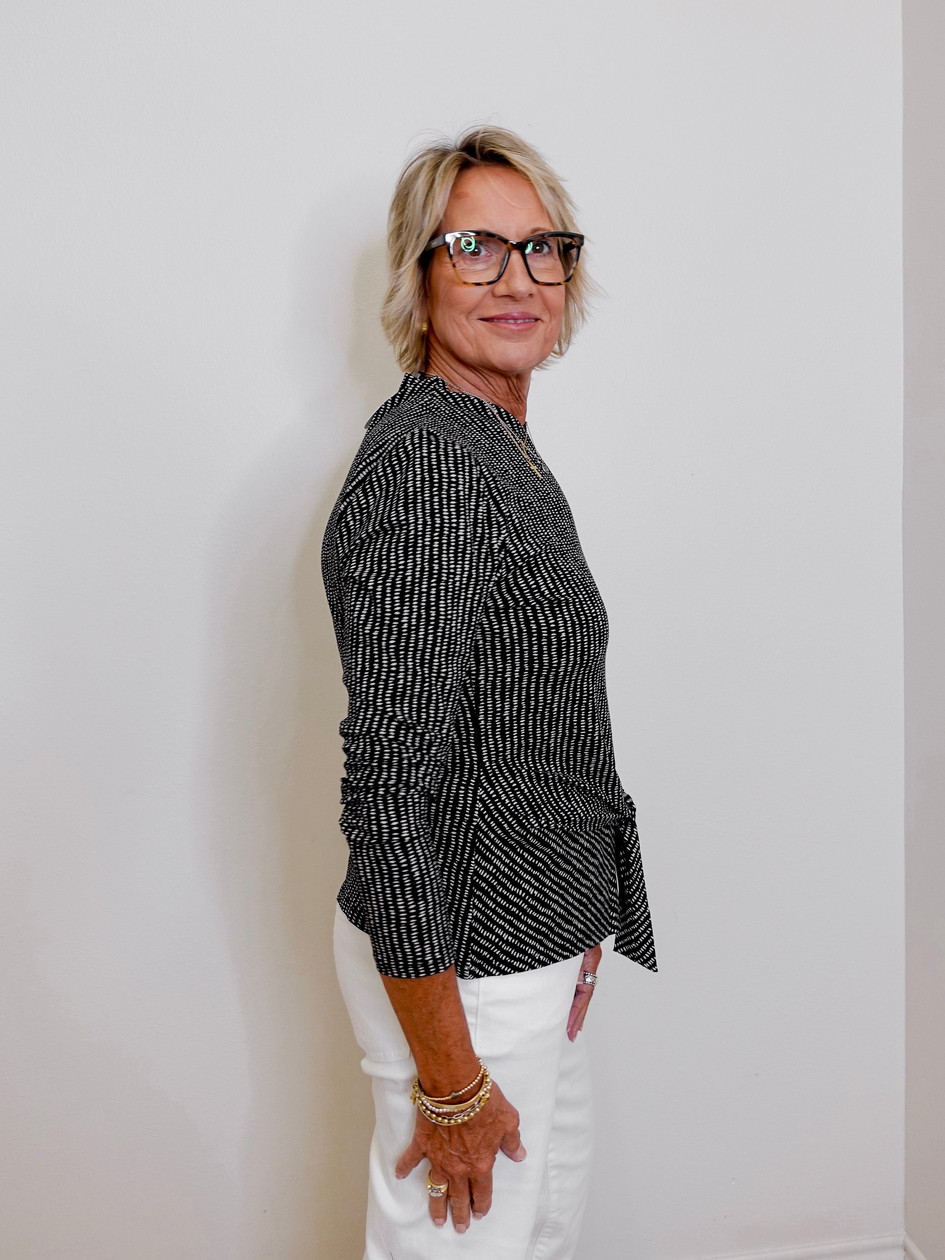Woman wearing a black patterned blouse and white pants against a plain background