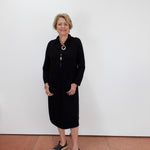 A woman with short blonde hair and glasses stands against a plain white wall wearing a black long-sleeve midi dress, a long silver-toned necklace with a tassel, and black slide sandals.