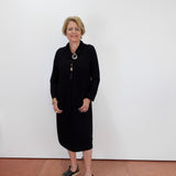 A woman with short blonde hair and glasses stands against a plain white wall wearing a black long-sleeve midi dress, a long silver-toned necklace with a tassel, and black slide sandals.