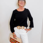 Woman wearing a black top and white pants, holding a floral clutch against a white wall.