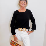 Woman wearing a black top and white pants, holding a floral clutch against a white wall.
