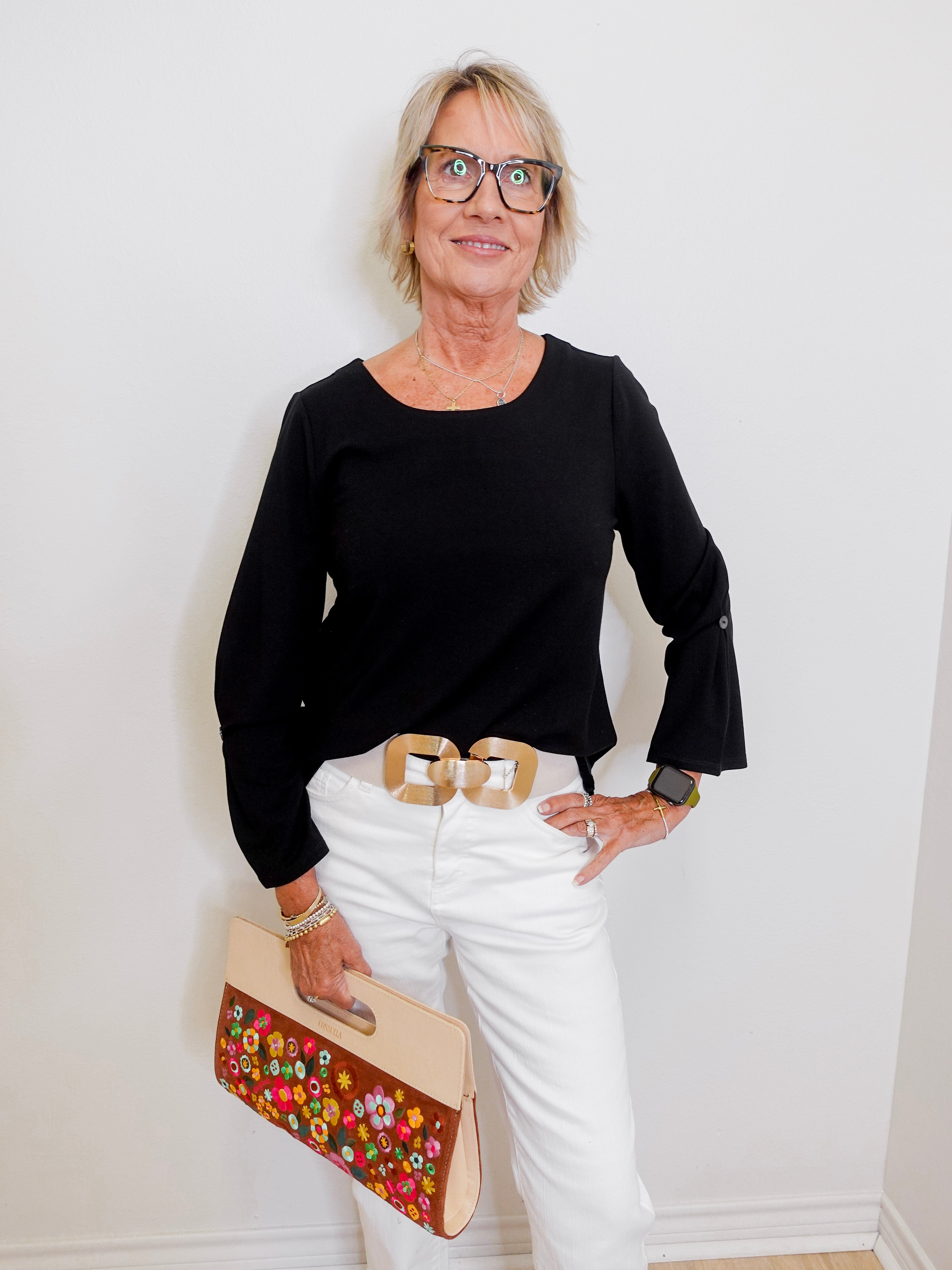 Woman wearing a black top and white pants, holding a floral clutch against a white wall.
