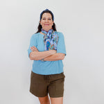 Woman wearing a blue shirt, green shorts, and a colorful scarf with arms crossed against a white background