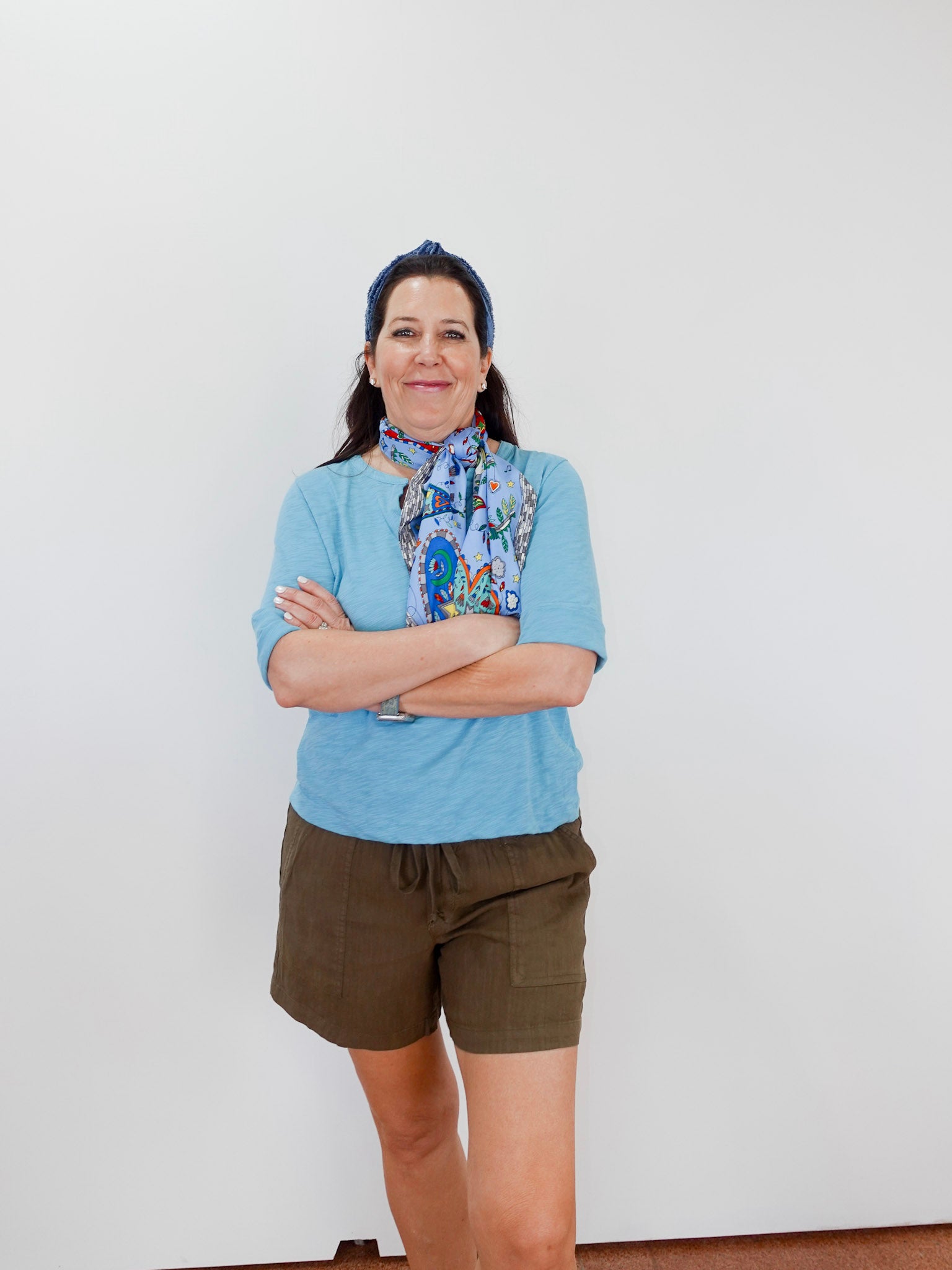 Woman wearing a blue shirt, green shorts, and a colorful scarf with arms crossed against a white background