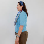 Woman wearing a blue shirt and green shorts with a colorful scarf, standing against a white wall.