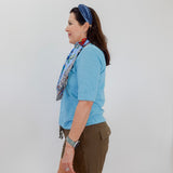Woman wearing a blue shirt and green shorts with a colorful scarf, standing against a white wall.