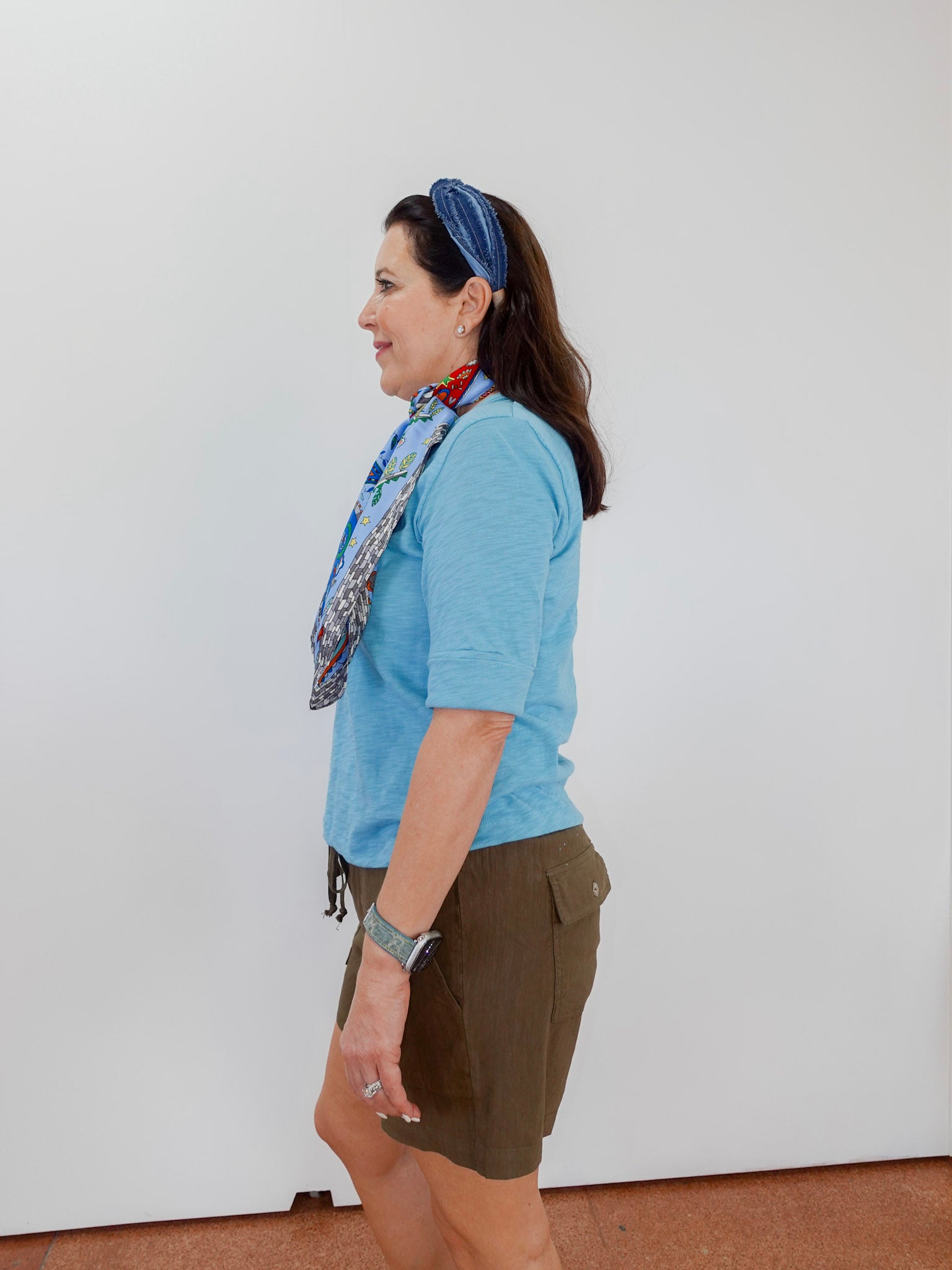 Woman wearing a blue shirt and green shorts with a colorful scarf, standing against a white wall.
