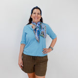 Woman wearing a blue shirt and brown shorts with a colorful scarf, standing against a white background.