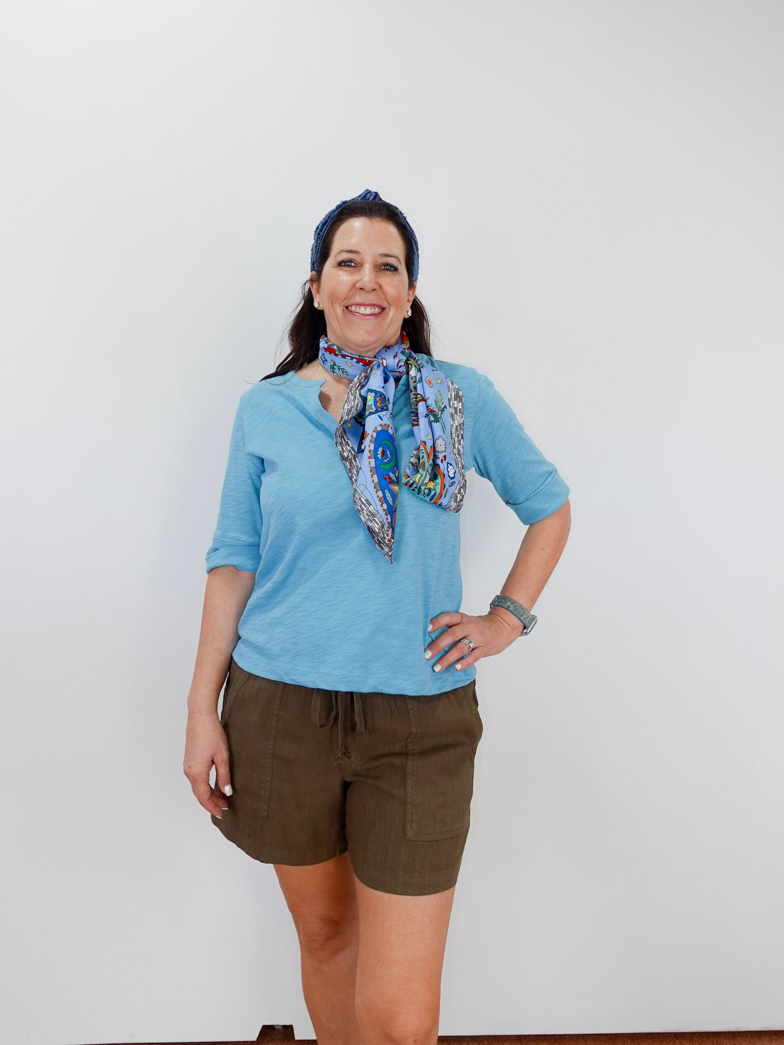 Woman wearing a blue shirt and brown shorts with a colorful scarf, standing against a white background.