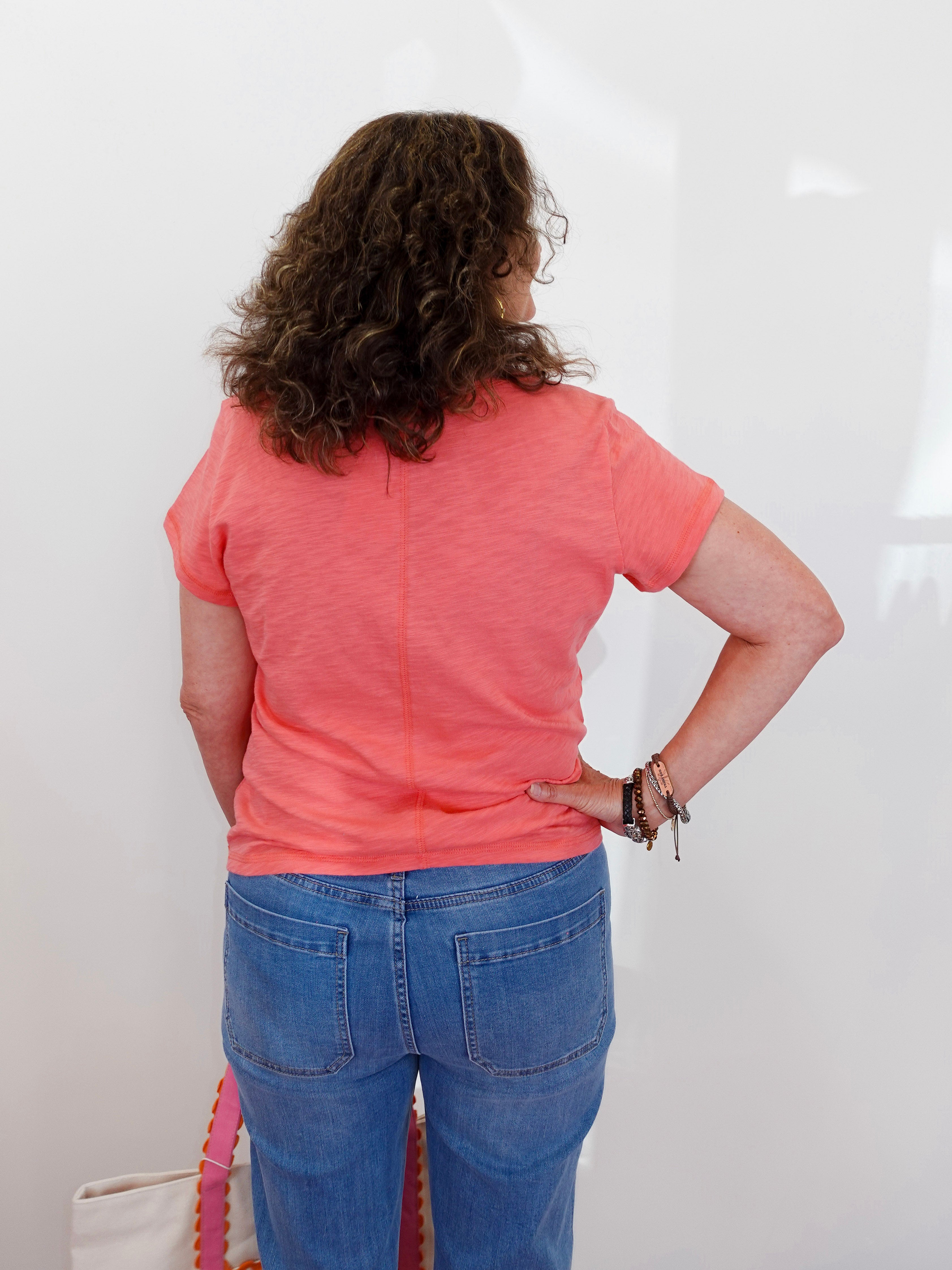 Person wearing a pink shirt and blue jeans standing against a white wall.
