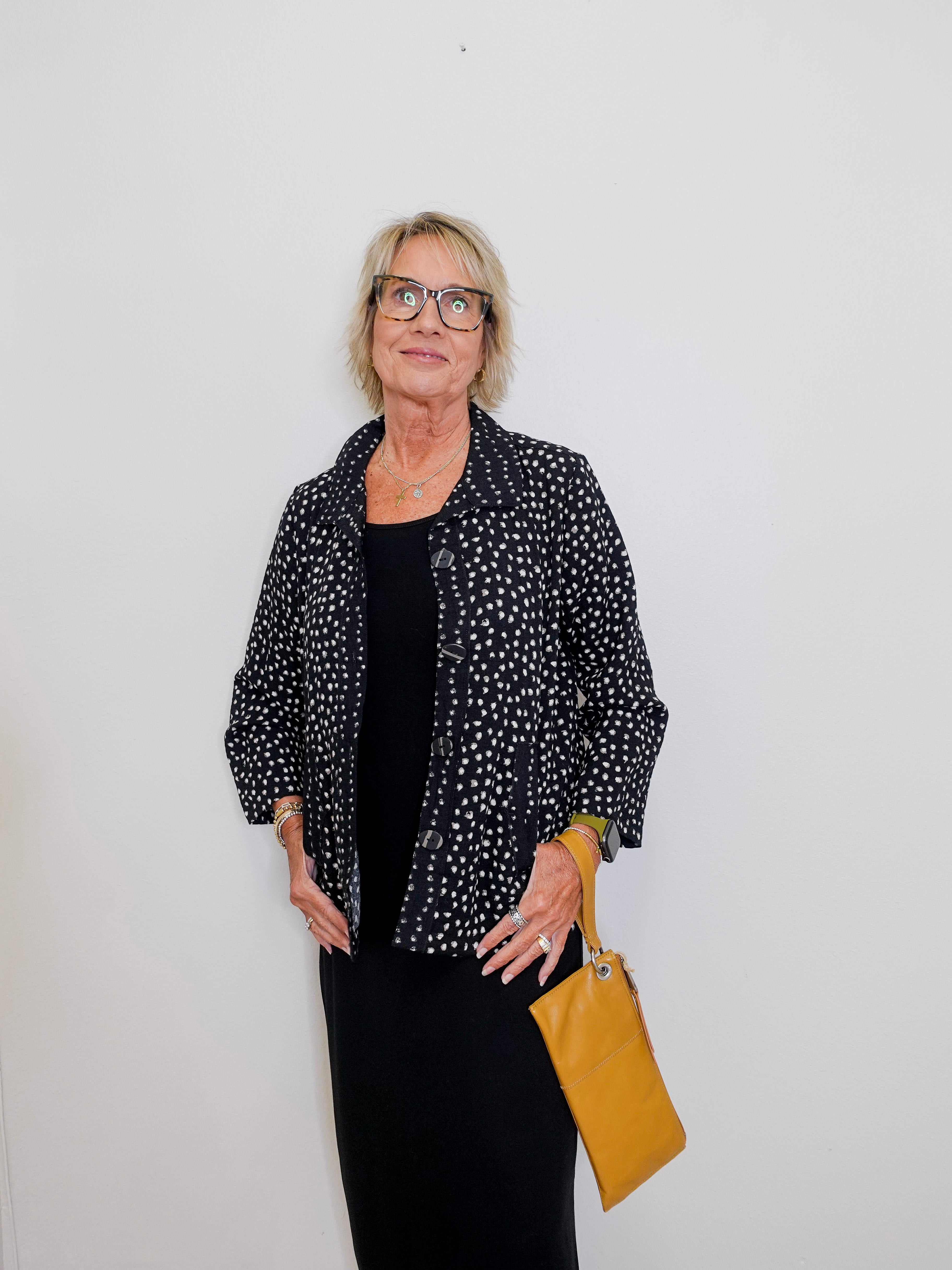 Woman wearing a black polka dot jacket over a black top, holding a yellow bag against a white background