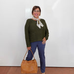 A woman stands against a white wall wearing a dark olive green knit sweater, navy blue pants, and beige heeled sandals, holding a tan leather tote bag. She is wearing a patterned cream scarf and glasses.