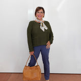A woman stands against a white wall wearing a dark olive green knit sweater, navy blue pants, and beige heeled sandals, holding a tan leather tote bag. She is wearing a patterned cream scarf and glasses.