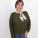 A woman wearing a textured olive green knit sweater, blue pants, and a patterned neck scarf, standing against a plain white background.