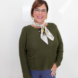 A woman wearing a textured olive green knit sweater, blue pants, and a patterned neck scarf, standing against a plain white background.