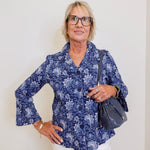 Woman wearing a blue floral blouse and white pants, holding a blue handbag against a plain background.