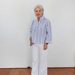 Woman wearing a blue and white striped shirt and white pants standing against a plain white wall.