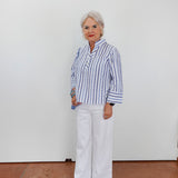 Woman wearing a blue and white striped shirt and white pants standing against a plain white wall.