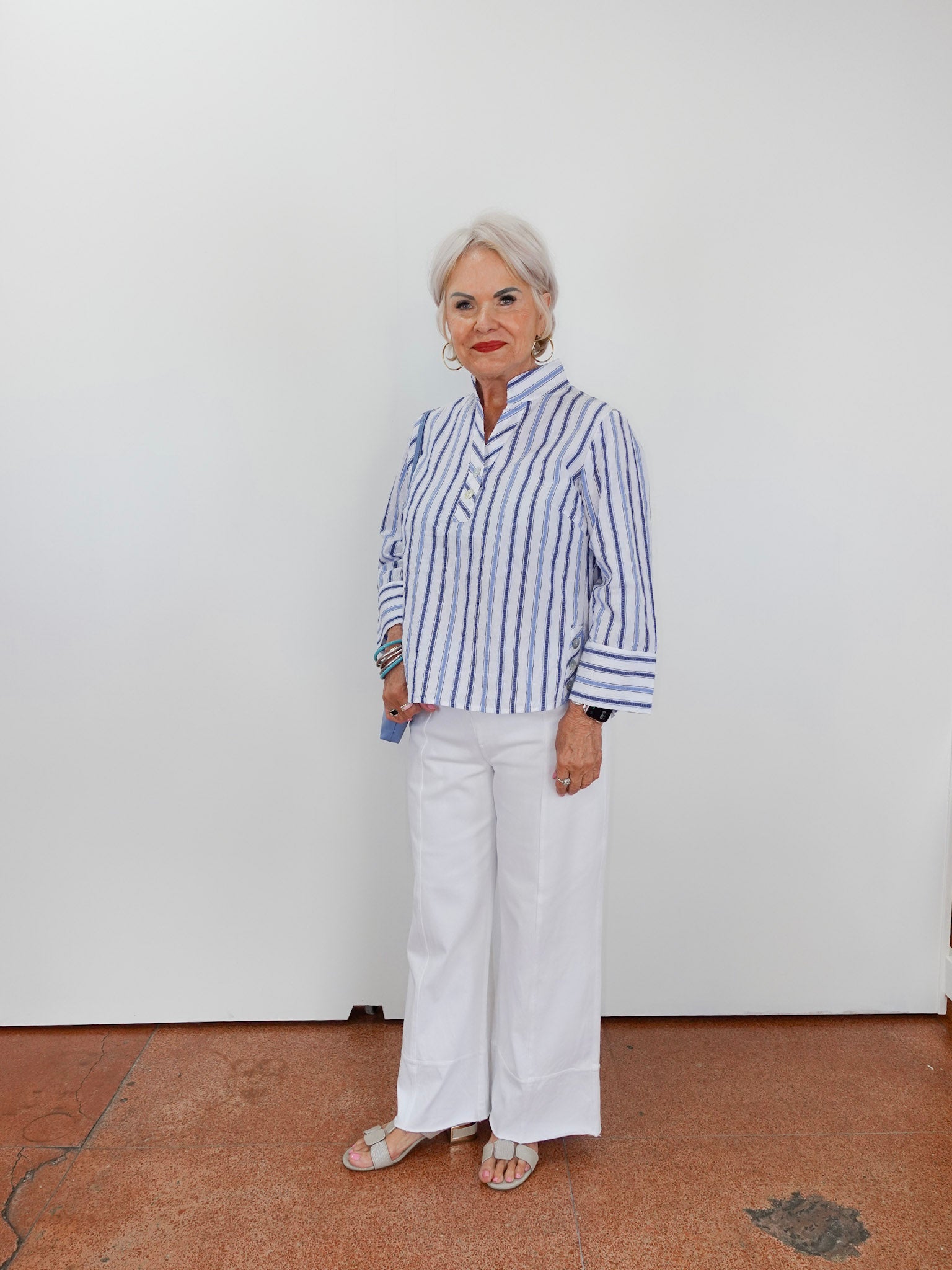 Woman wearing a blue and white striped shirt and white pants standing against a plain white wall.