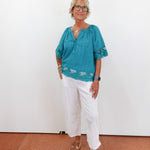 Woman wearing a teal blouse with white pants against a white wall.