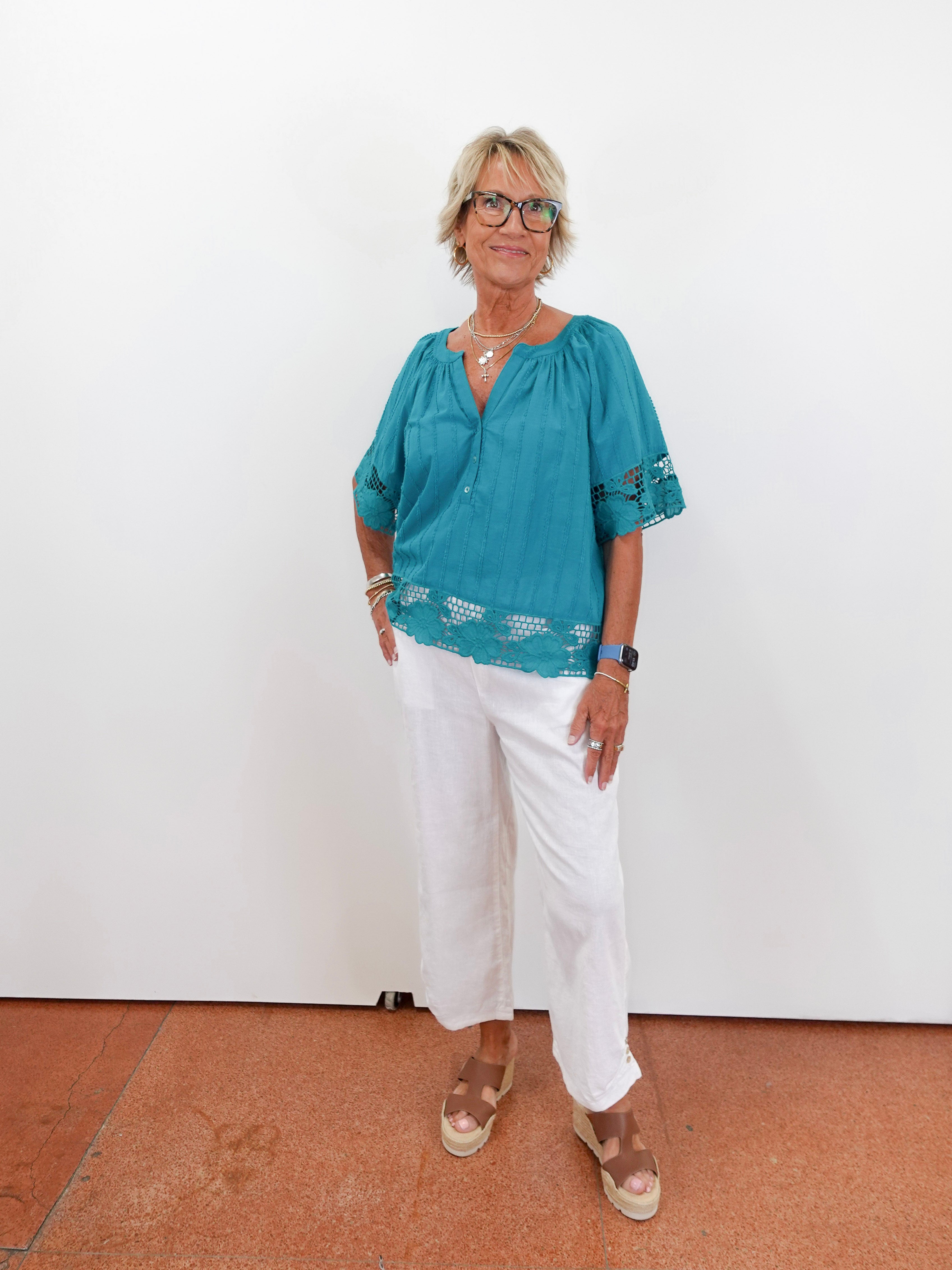 Woman wearing a teal blouse with white pants against a white wall.