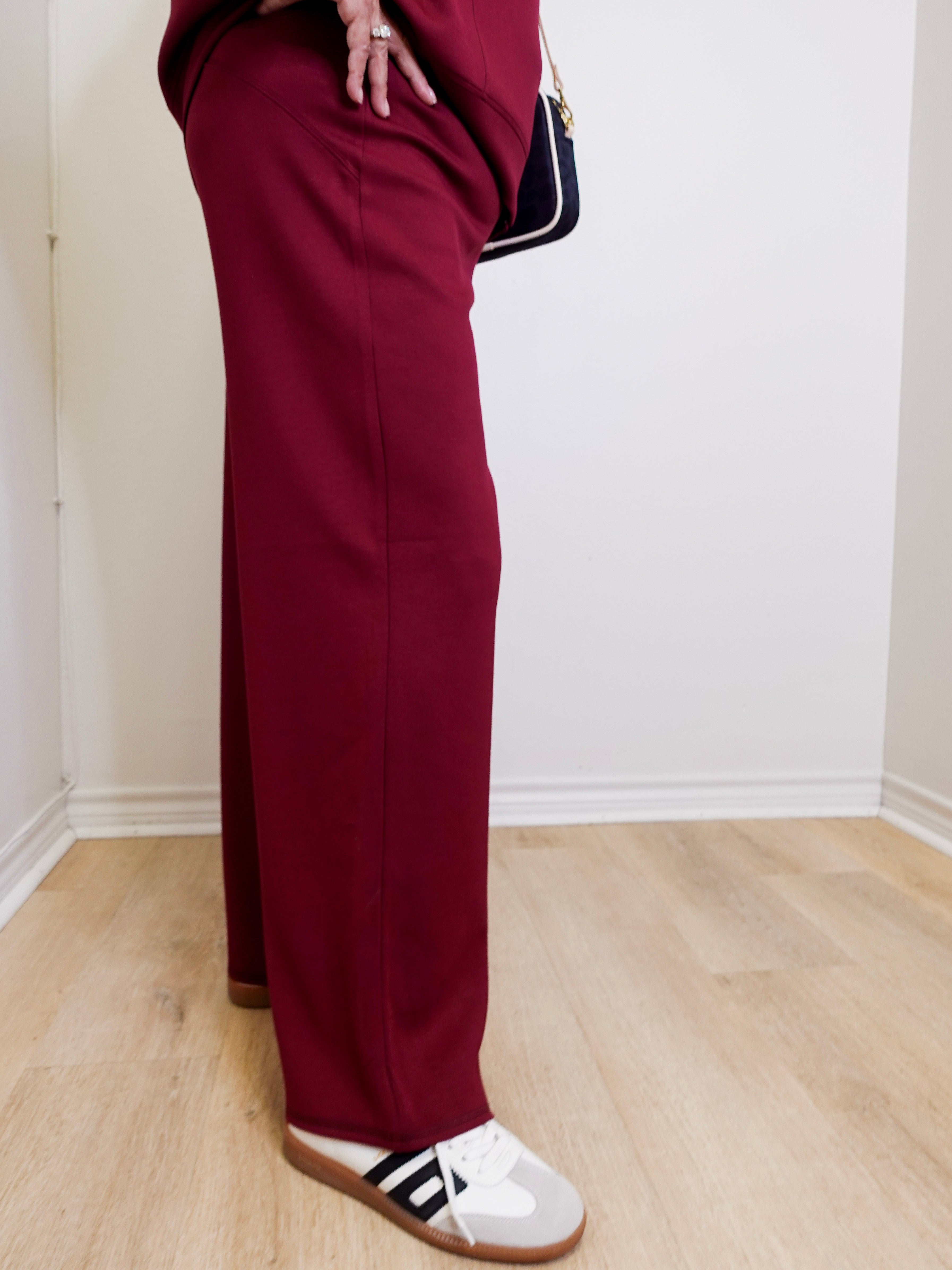 Person wearing burgundy pants and white sneakers with black stripes on a light wooden floor.