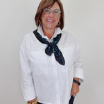 A woman with short brown hair and glasses, wearing a white button-down shirt and a dark blue patterned neck scarf, standing against a plain light gray background.
