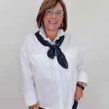 A woman with short brown hair and glasses, wearing a white button-down shirt and a dark blue patterned neck scarf, standing against a plain light gray background.