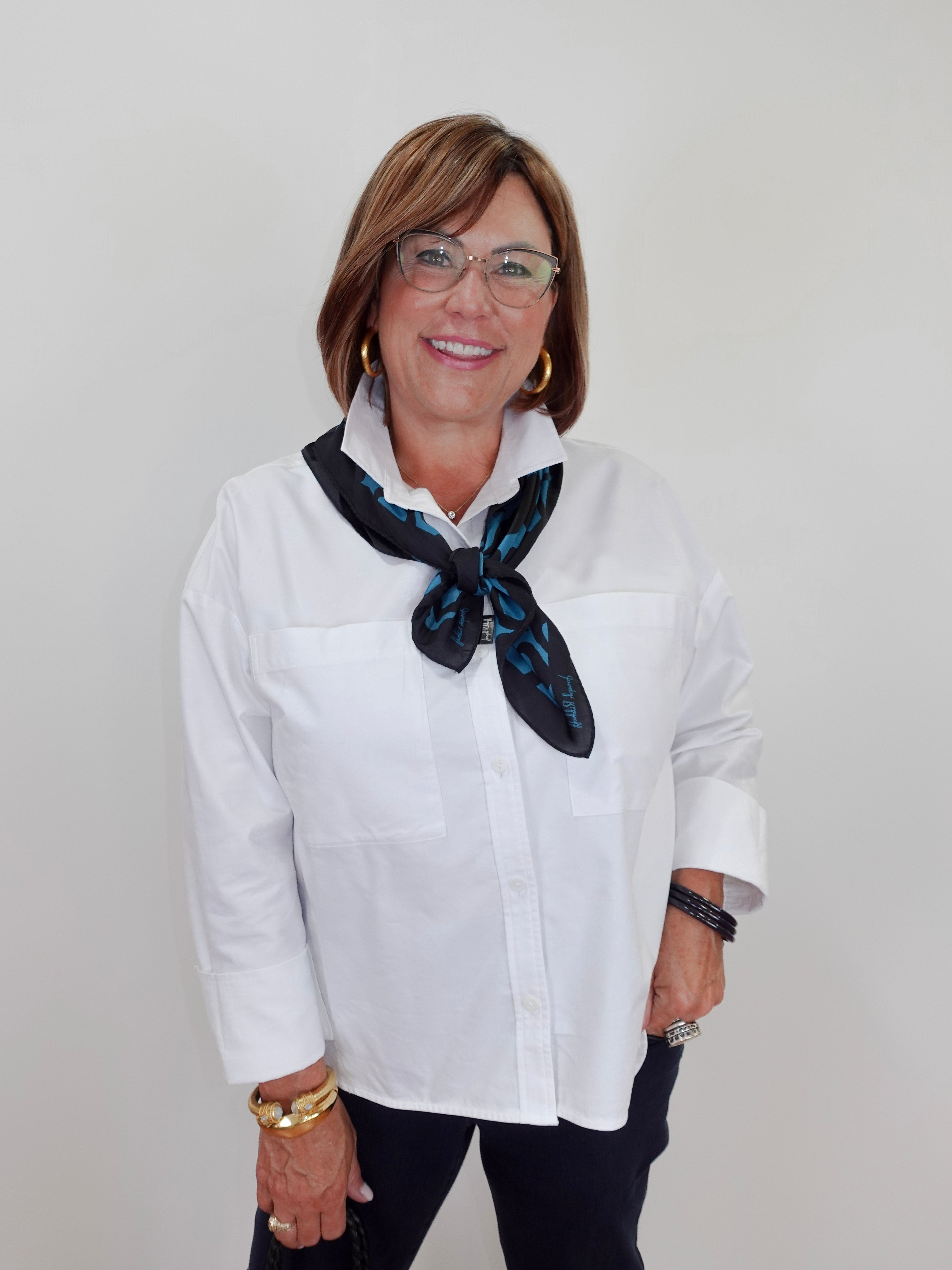 A woman with short brown hair and glasses, wearing a white button-down shirt and a dark blue patterned neck scarf, standing against a plain light gray background.