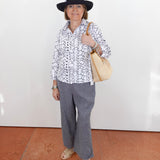 Woman wearing a black hat, patterned shirt, gray pants, and holding a beige handbag against a white wall.