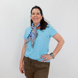 Woman wearing a light blue shirt and brown shorts with a patterned scarf, standing against a white background.
