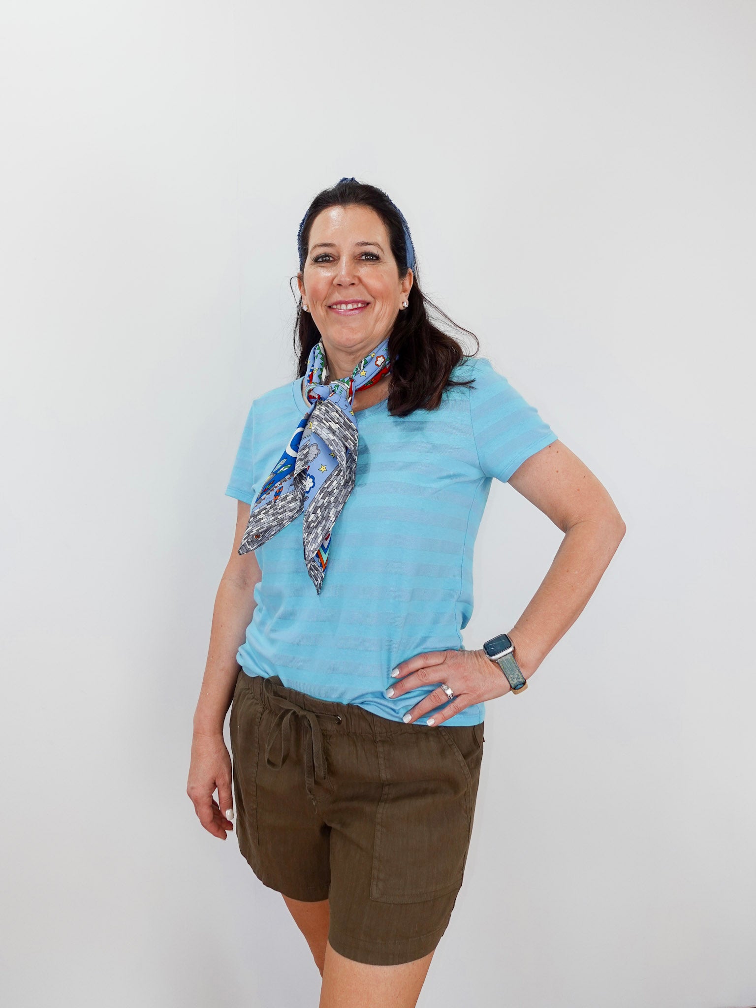 Woman wearing a light blue shirt and brown shorts with a patterned scarf, standing against a white background.