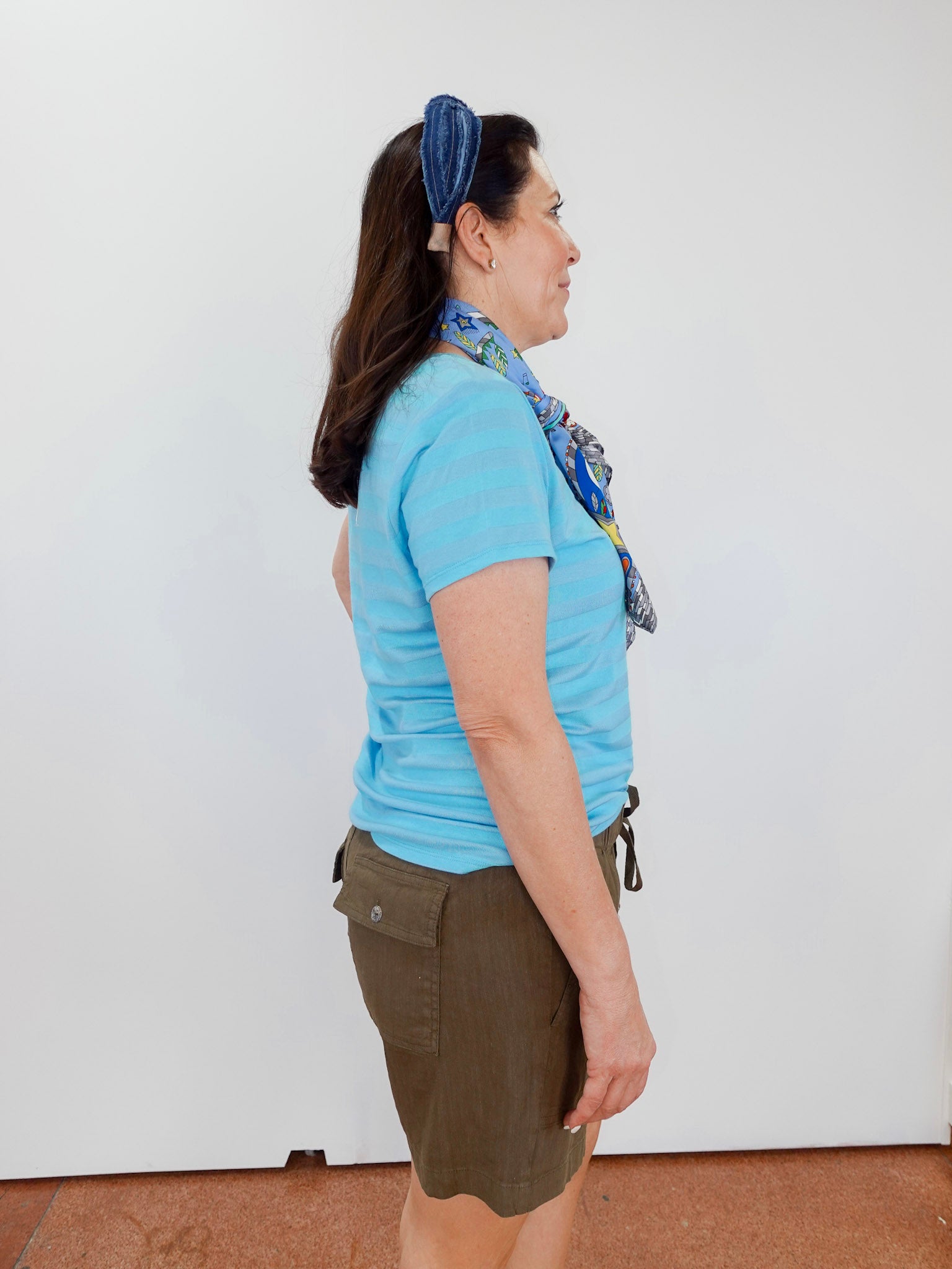 Woman wearing a blue shirt and brown shorts with a headband, standing against a white wall.